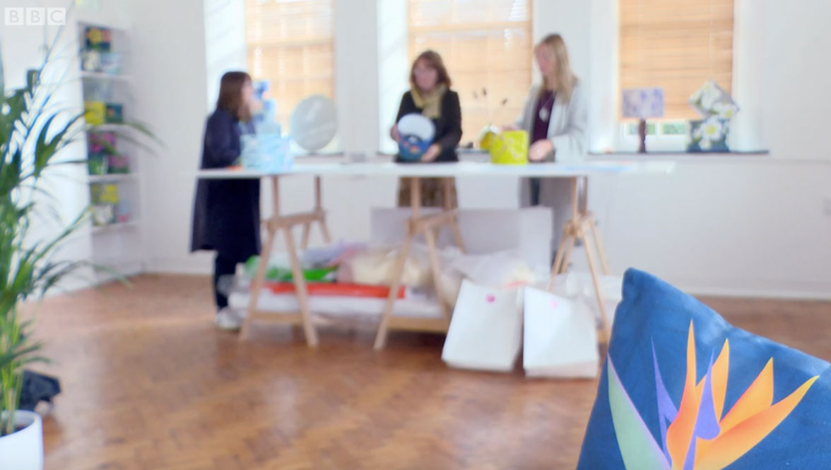 In the foreground the image shows an edge of a Bird of Paradise Flower cushion, and in the background out of focus Alison Bick is teaching how to make lampshades with her fabric designs.
